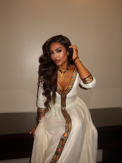 A woman wearing a white long-sleeve maxi traditional clothing  with colorful geometric embroidery along the neckline, front opening, and cuffs, posed against a plain beige wall.