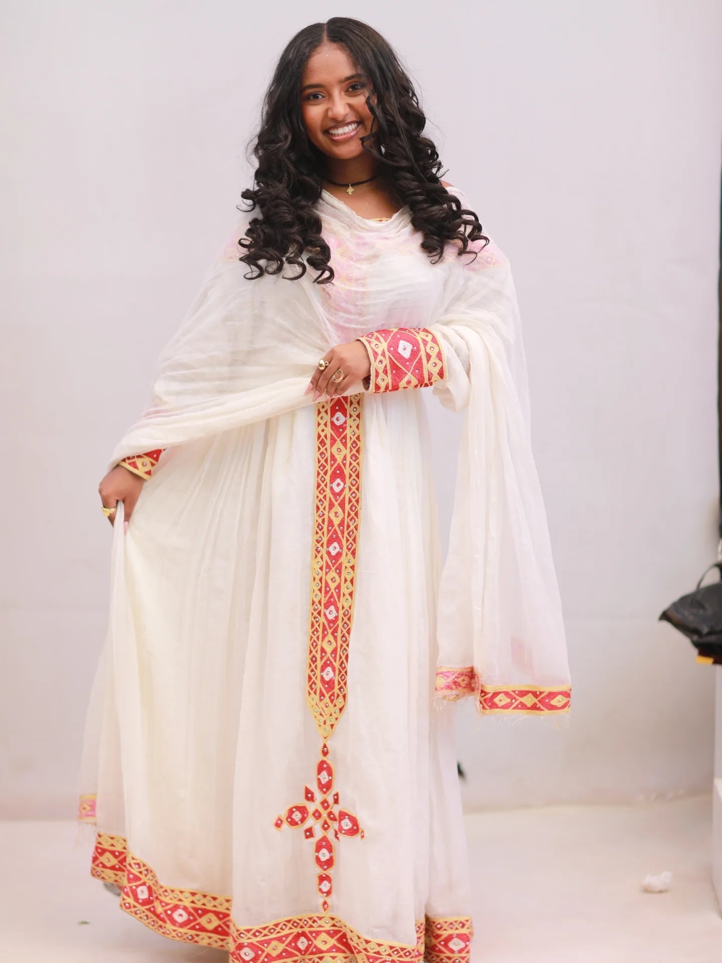 A woman modeling a traditional white Ethiopian Dress with red and gold geometric embroidery, featuring a matching sheer dupatta.