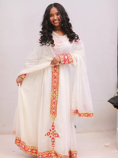 A woman modeling a traditional white Ethiopian Dress with red and gold geometric embroidery, featuring a matching sheer dupatta.