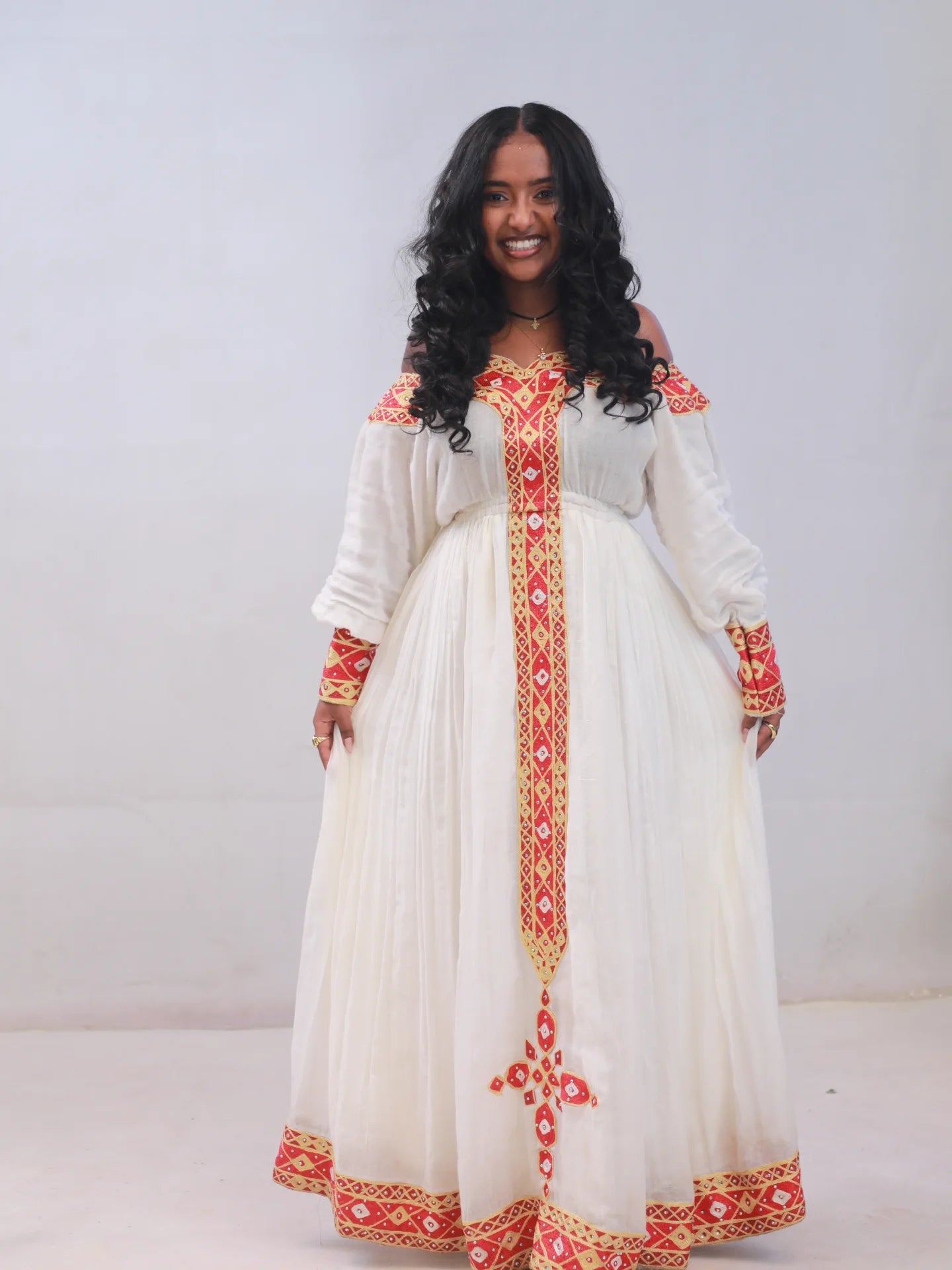 A woman modeling a long off-the-shoulder Ethiopian Dress with red and gold embroidered details, standing against a plain light gray studio background.