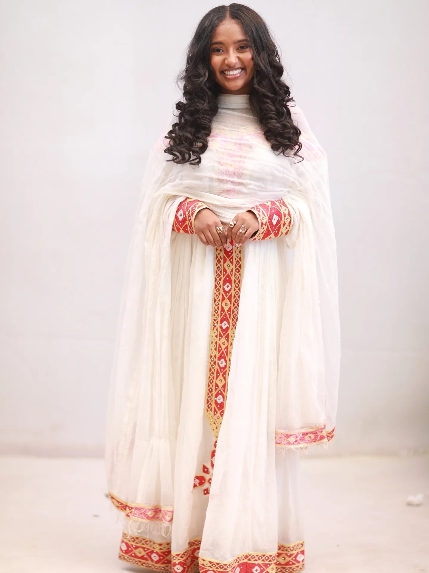 Woman wearing a white traditional Ethiopian Dress with red and gold patterns on a plain background