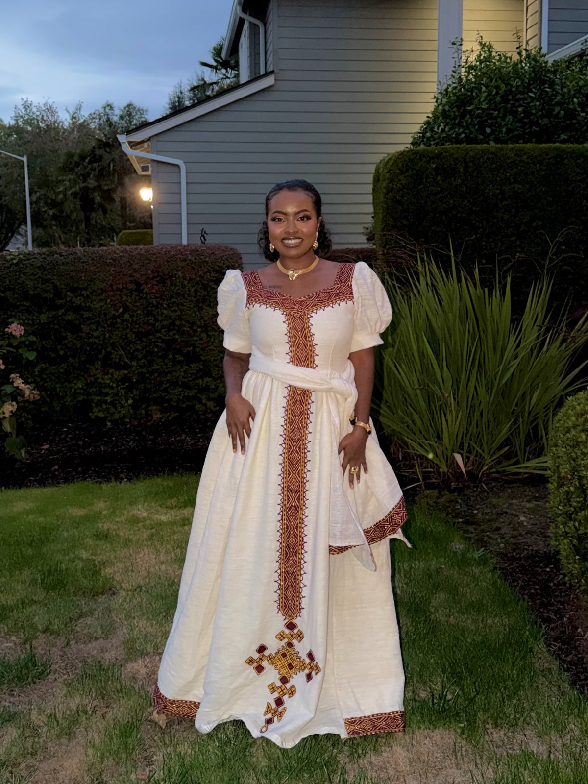 A woman stands outdoors in a grassy yard wearing a long cream Ethiopian dress with brown and gold embroidered details, puff sleeves, and a matching waist tie.