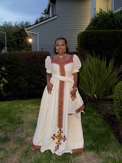 A woman stands outdoors in a grassy yard wearing a long cream Ethiopian dress with brown and gold embroidered details, puff sleeves, and a matching waist tie.