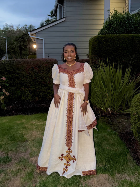 A woman stands outdoors in a grassy yard wearing a long cream Ethiopian dress with brown and gold embroidered details, puff sleeves, and a matching waist tie.
