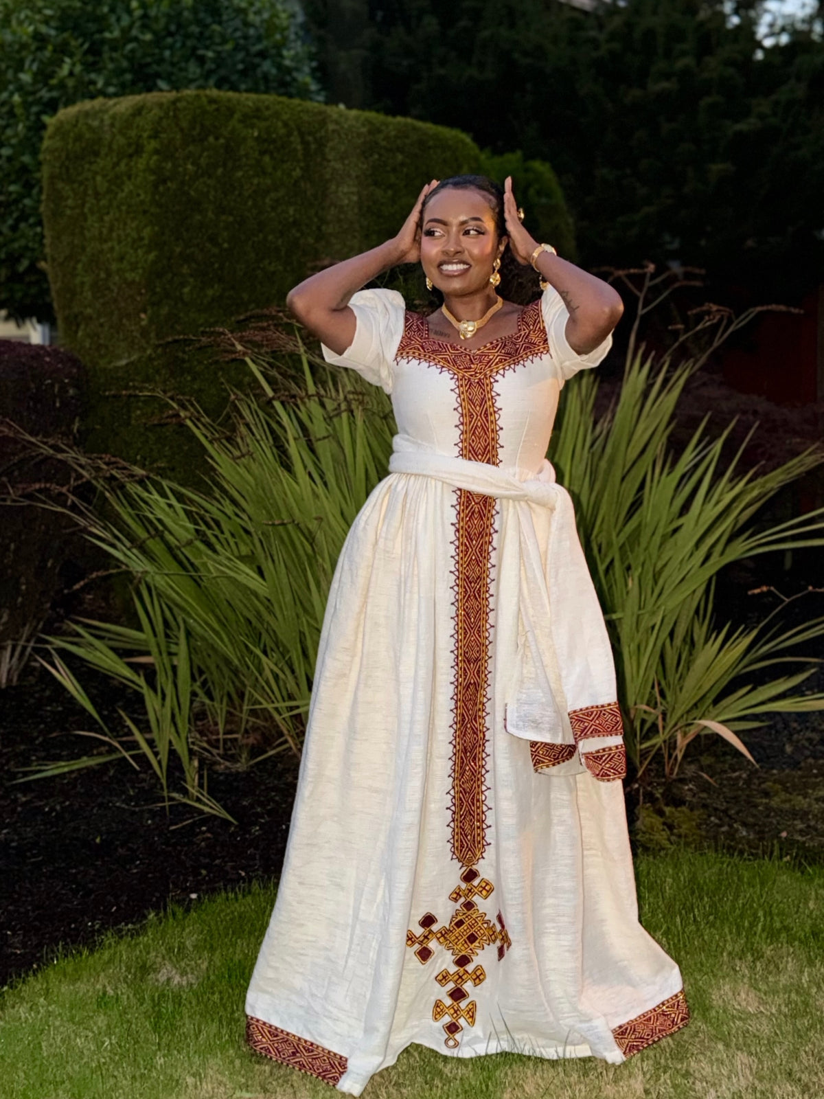 A woman wearing a long white traditional Ethiopian dress with brown and gold embroidered details, standing outdoors in a garden setting.