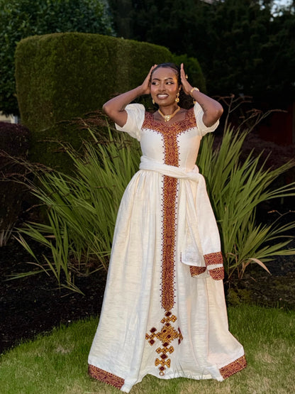 A woman wearing a long white traditional Ethiopian dress with brown and gold embroidered details, standing outdoors in a garden setting.