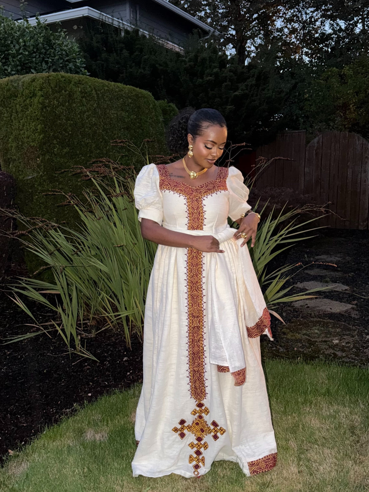 A woman wearing a long white Ethiopian dress with ornate brown and gold embroidered details, standing outdoors in a garden setting.