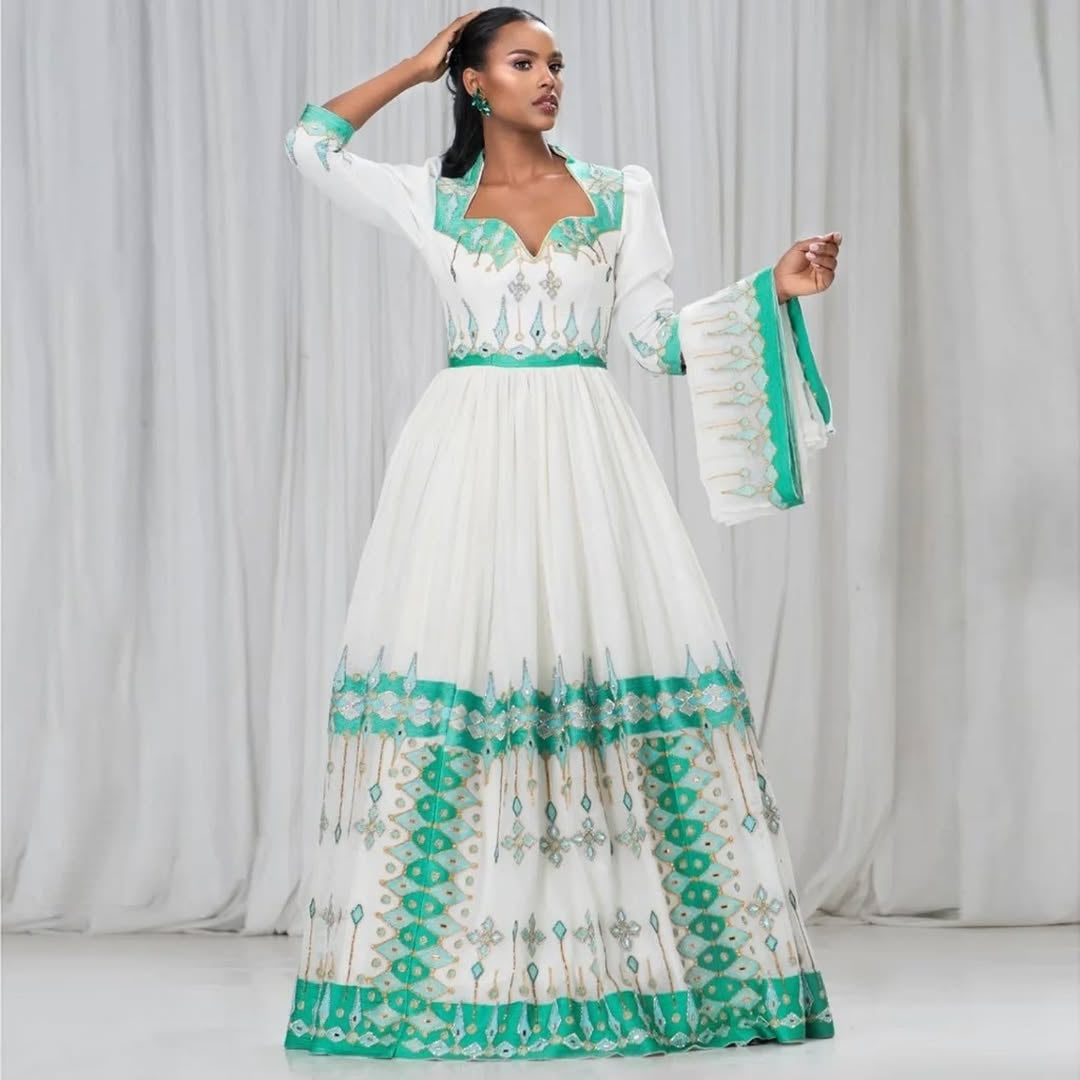 Woman wearing a white and green Ethiopian Kemis with intricate patterns against a plain background