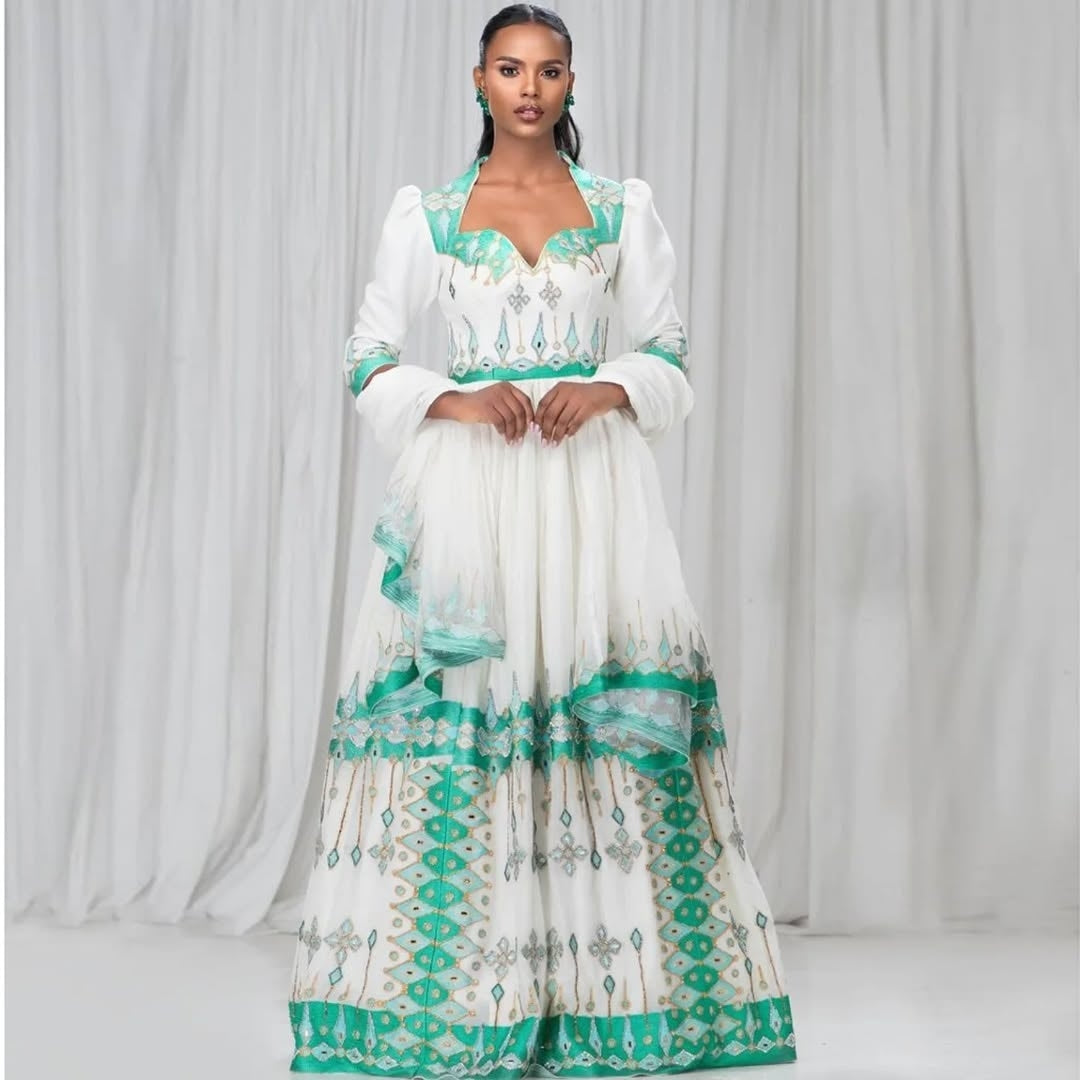 Woman wearing a white and green embroidered Ethiopian Kemis against a white curtain background