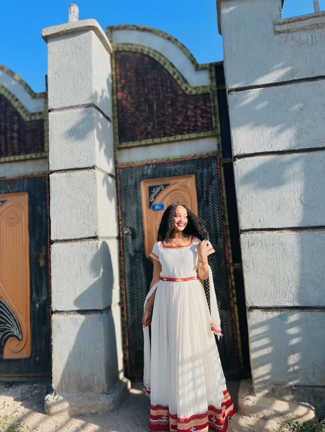 Woman in a Ethiopian dress with red accents standing in front of a decorative gate.