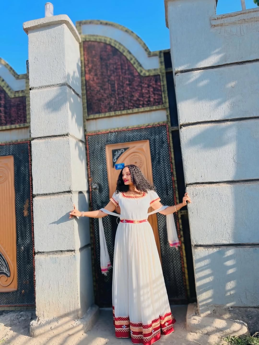 Woman in a white Ethiopian dress with red accents standing in front of a decorative wall.