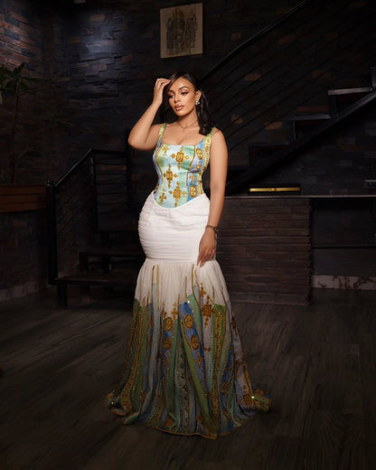 A woman models a sleeveless Habesha kemis with a light blue and gold patterned bodice, a fitted white waist, and a flowing skirt featuring blue, green, and gold vertical patterns, set against a dark wooden staircase background.
