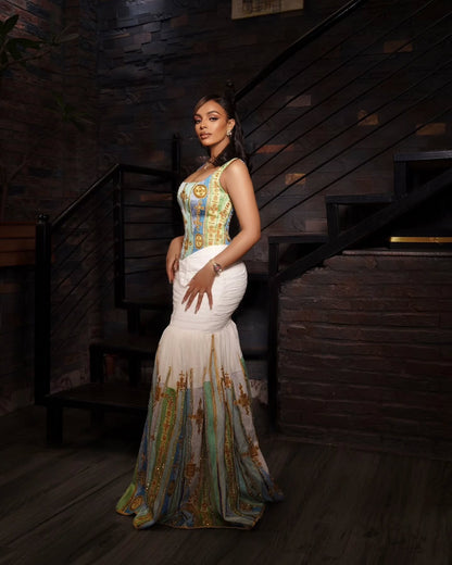 A woman models a sleeveless mermaid-style evening Habesha kemis in a dimly lit indoor setting with a staircase and brick wall. The dress has a fitted bodice with blue, gold, and green ornamental patterning, a white ruched waist, and a flowing skirt with a matching multicolor pattern.
