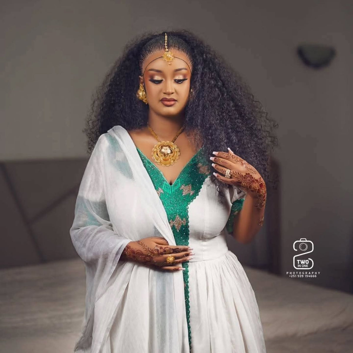 A woman with long, curly black hair models a traditional white and green Ethiopian Traditional Clothing with gold jewelry, including a prominent gold necklace and a gold headpiece. She has henna on her hands and is posed in a softly lit indoor setting.
