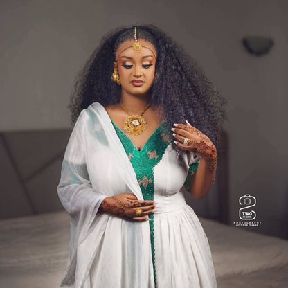 A woman with long, curly black hair models a traditional white and green Ethiopian Traditional Clothing with gold jewelry, including a prominent gold necklace and a gold headpiece. She has henna on her hands and is posed in a softly lit indoor setting.