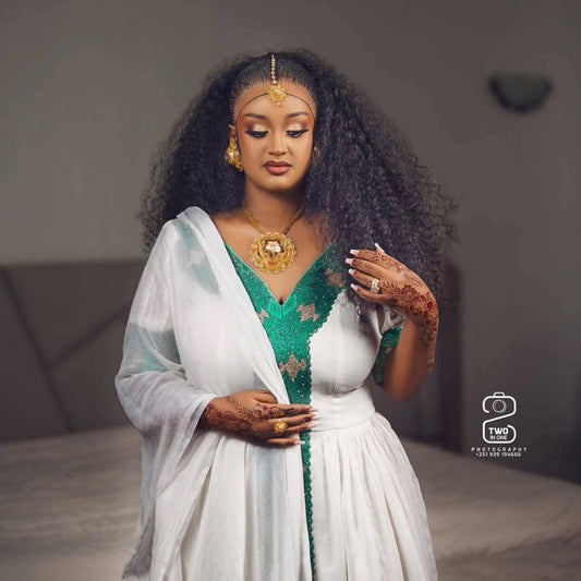 A woman with long, curly black hair models a traditional white and green Ethiopian Traditional Clothing with gold jewelry, including a prominent gold necklace and a gold headpiece. She has henna on her hands and is posed in a softly lit indoor setting.