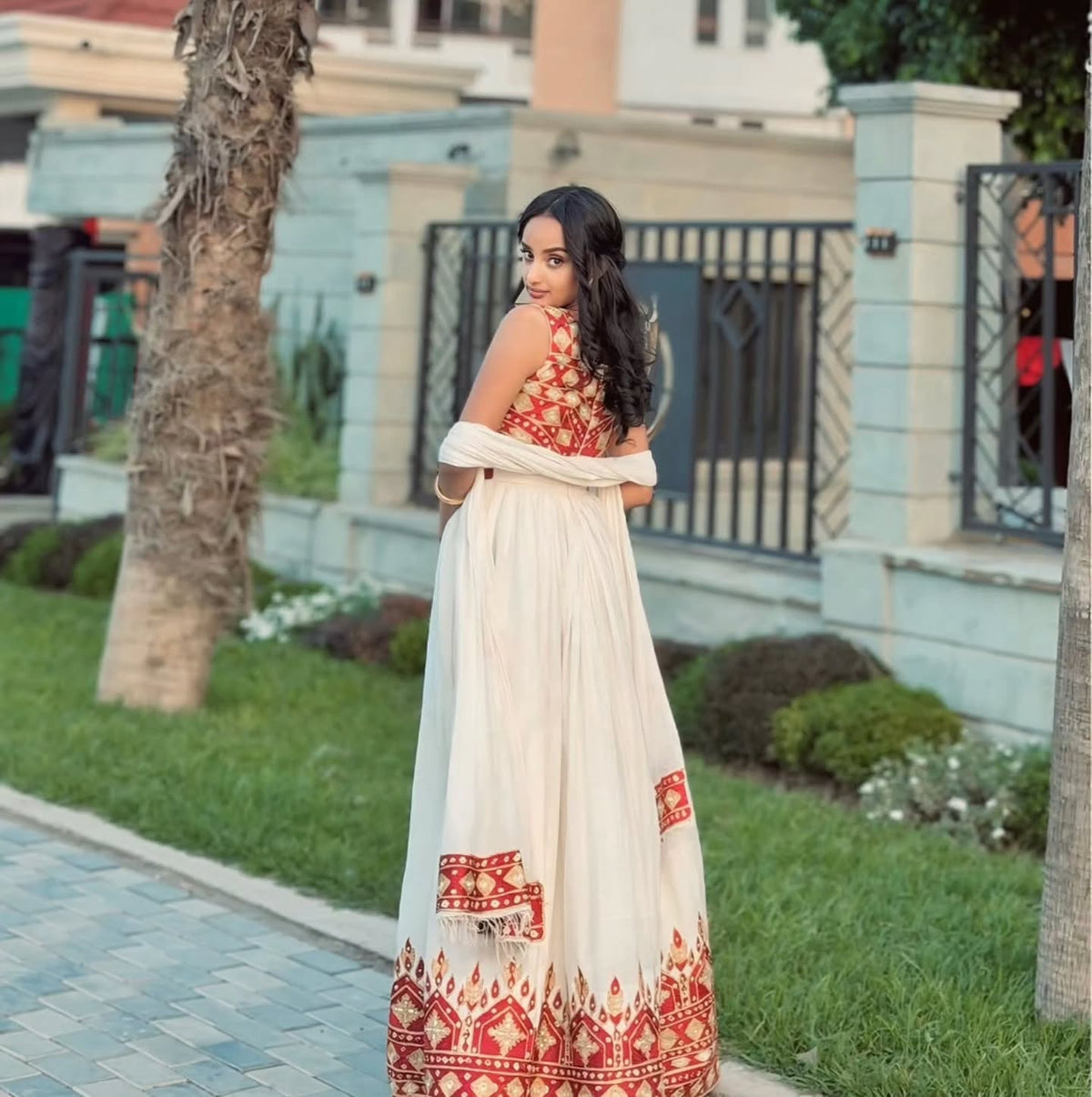 A woman wearing a traditional Ethiopian National Dresses with a cream-colored flared skirt and a red embroidered halter top, standing outdoors on a paved path with grass and trees in the background.