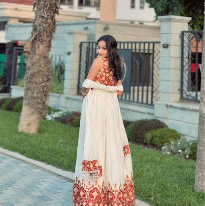 A woman wearing a traditional Ethiopian National Dresses with a cream-colored flared skirt and a red embroidered halter top, standing outdoors on a paved path with grass and trees in the background.
