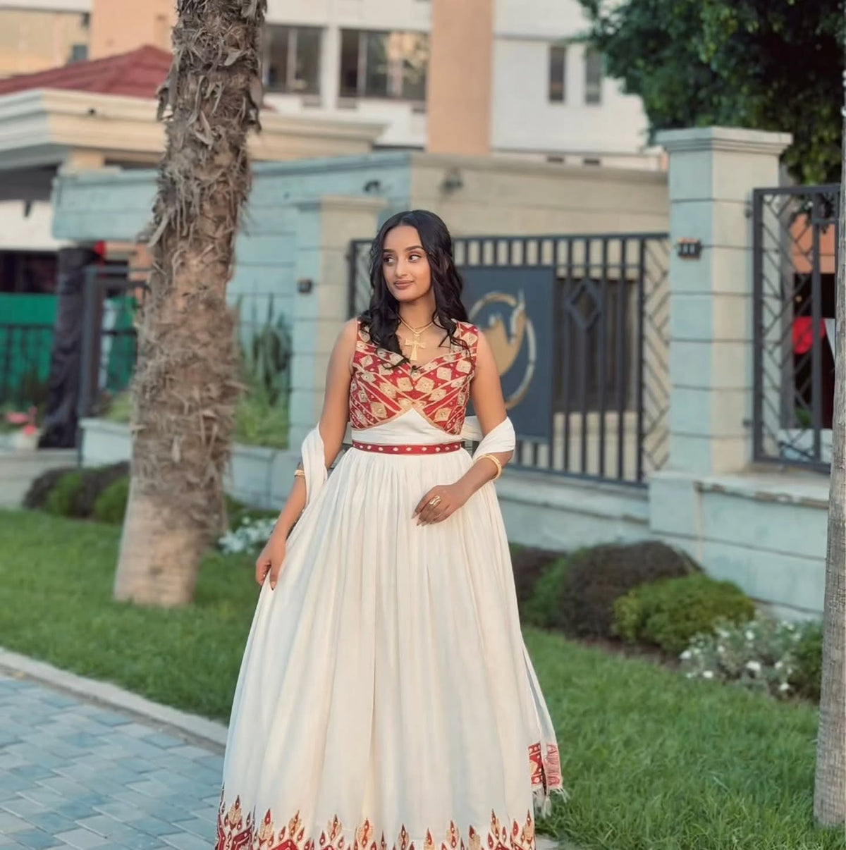 A woman wearing a traditional Ethiopian National Dresses with a red and gold patterned sleeveless bodice and a long cream skirt with red border detailing, standing outdoors in front of a light blue building and greenery.