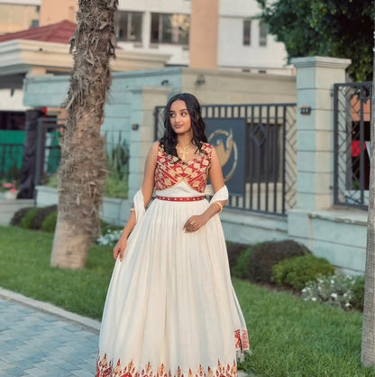 A woman wearing a traditional Ethiopian National Dresses with a red and gold patterned sleeveless bodice and a long cream skirt with red border detailing, standing outdoors in front of a light blue building and greenery.