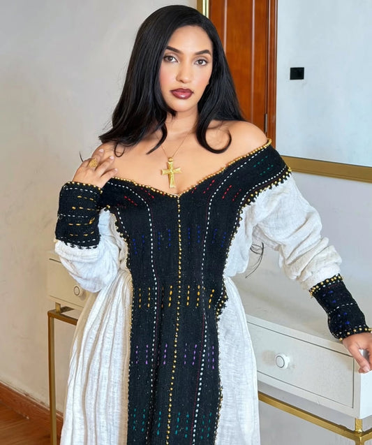 A woman wearing a black off-the-shoulder Habesha Dress  with white long sleeves and colorful embroidered details, standing indoors in front of a white dresser.