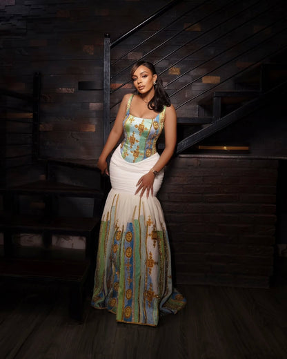 A woman stands in a dimly lit interior with dark wood paneling and stairs, wearing a sleeveless mermaid-style Habesha kemis with a turquoise and gold patterned bodice and skirt panels, and a white gathered waist section.