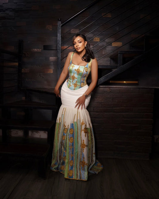 A woman stands in a dimly lit interior with dark wood paneling and stairs, wearing a sleeveless mermaid-style Habesha kemis with a turquoise and gold patterned bodice and skirt panels, and a white gathered waist section.