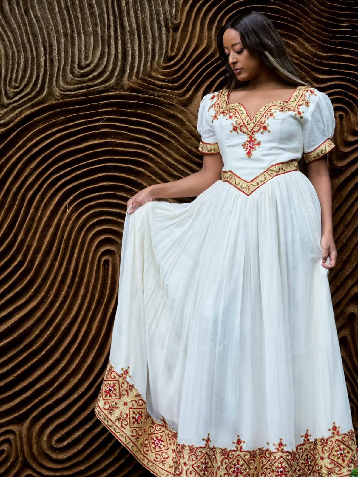 A woman wearing a white traditional-style Ethiopian Dress  with red and gold embroidery, standing against a textured brown background.