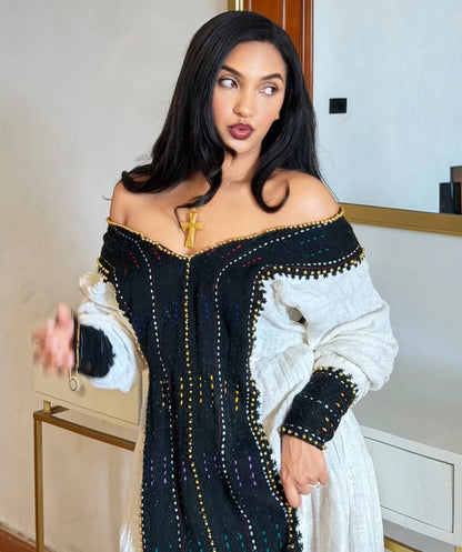 A woman wearing a black off-the-shoulder Habesha Dress  with colorful embroidered details and a white textured long-sleeve layer, posed indoors against a light-colored wall.