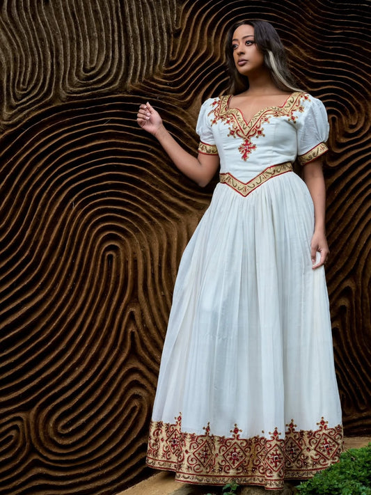 A woman wearing a long white Ethiopian Dress  with ornate red and gold embroidery, standing in front of a textured brown background.
