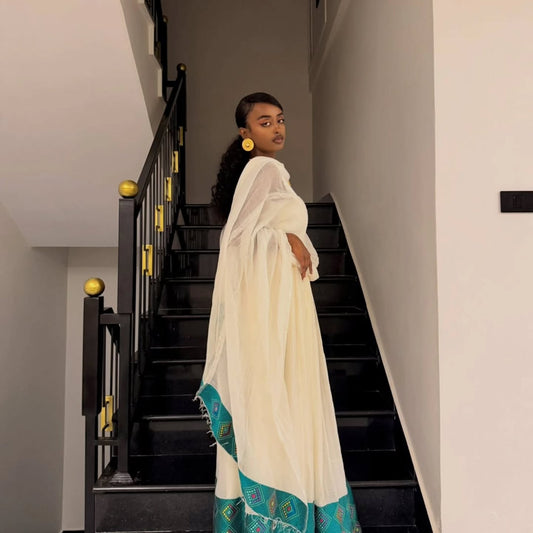 A woman wearing a white traditional ethiopian dress for women with a teal patterned border stands on a black staircase in an indoor setting.