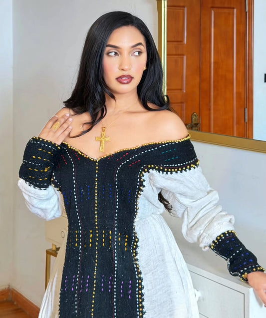 A woman wearing a black and white off-the-shoulder Habesha Dress  with colorful embroidered details, standing indoors in front of a mirror and wooden door.
