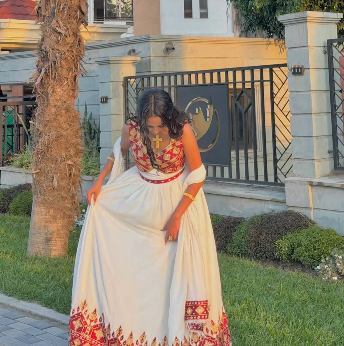 A woman wearing a traditional Ethiopian National Dresses with a white skirt, red and gold embroidered bodice, and a matching white dupatta, standing outdoors in front of a fence and palm tree.