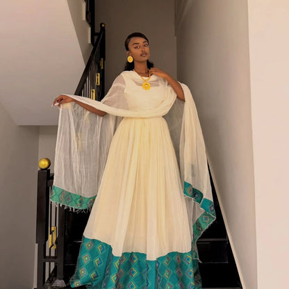 A woman stands on an indoor staircase wearing a traditional ethiopian dress for women with a cream-colored dress and dupatta featuring teal borders with geometric patterns, accessorized with gold jewelry.