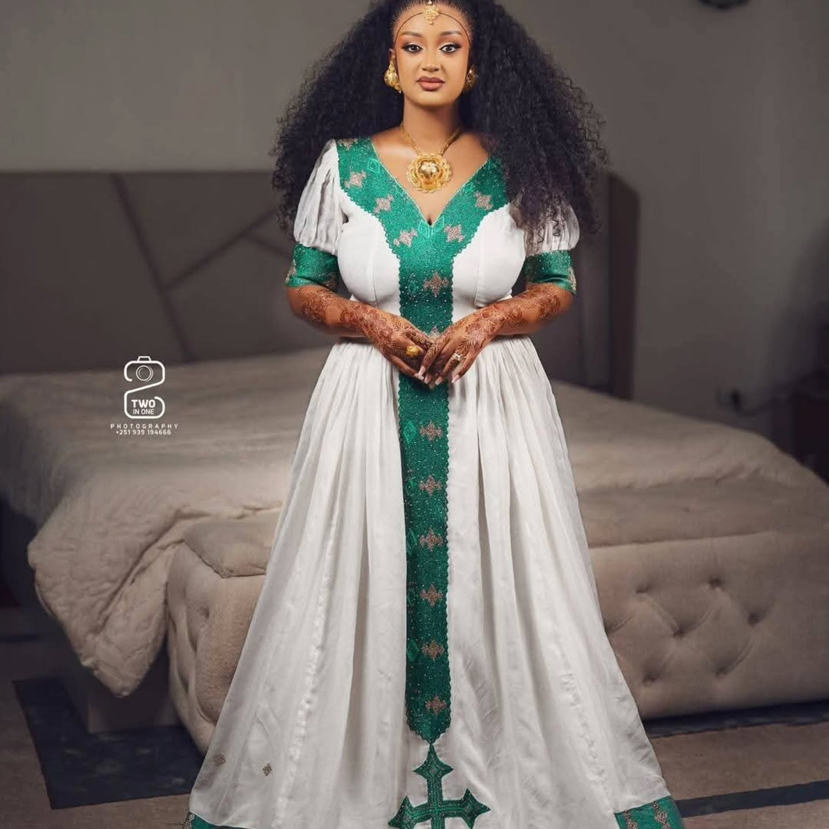 A woman stands in a bedroom wearing a long white Ethiopian Traditional Clothing with green embroidered trim and puff sleeves. The dress has a deep V-neckline and a vertical green embroidered panel down the front.
