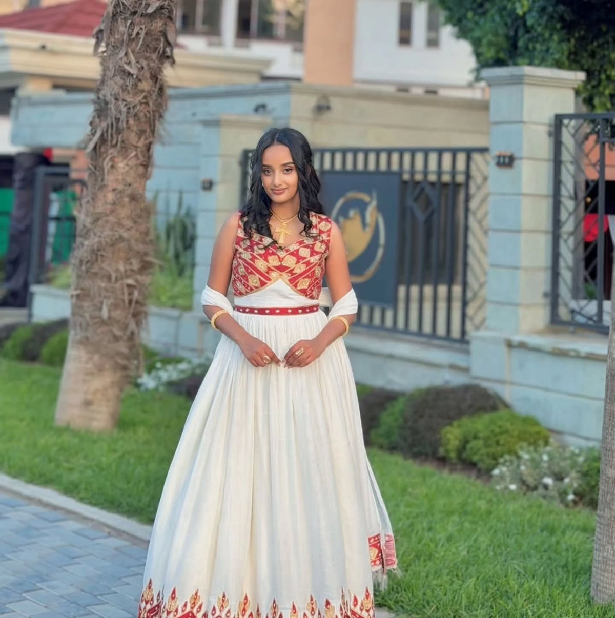 A woman stands outdoors wearing a traditional Ethiopian National Dresses with a red and gold patterned bodice and a red waistband, with white sleeves. The background shows a residential area with a gate, trees, and grass.
