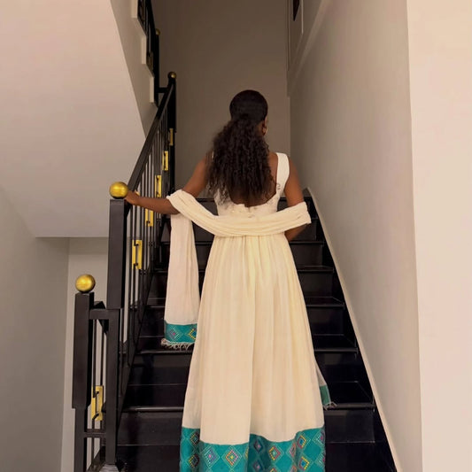 A woman viewed from behind wearing a cream-colored ethiopian dress for women with a teal and gold patterned hem, standing on a black staircase indoors.