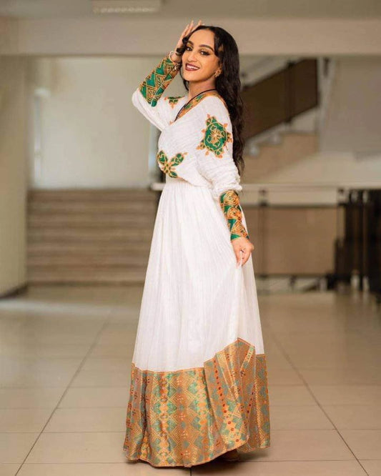 Woman in a white habesha dress with green and orange patterns posing indoors.
