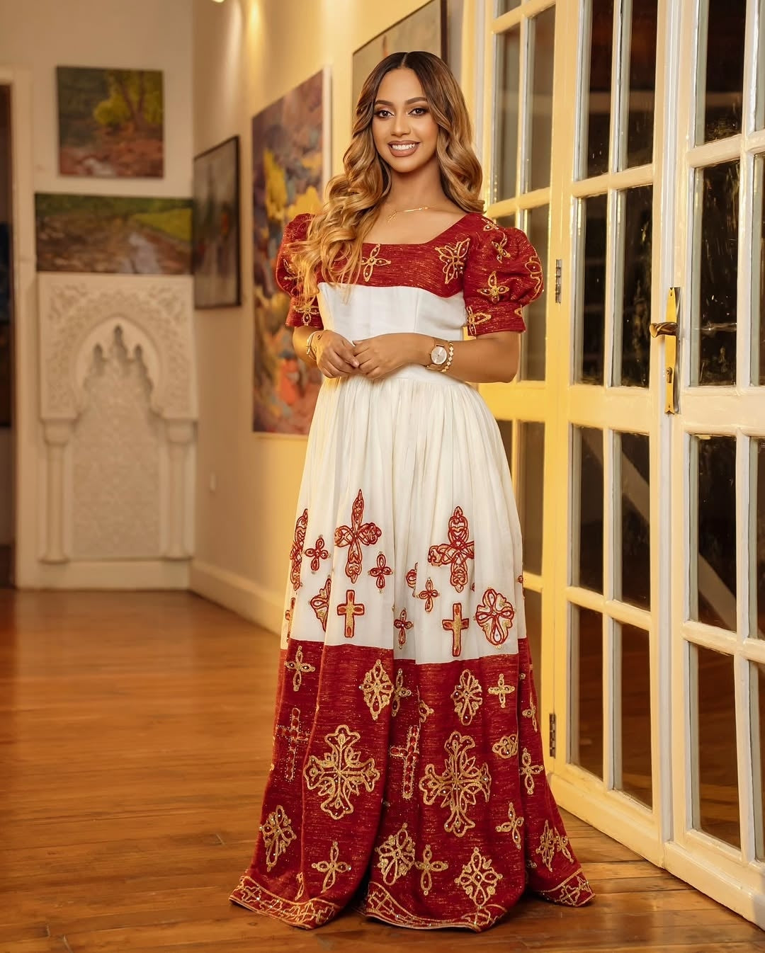 Woman wearing a red and white embroidered Ethiopian cultural dress in an indoor setting with wooden floor and decorative wall art.