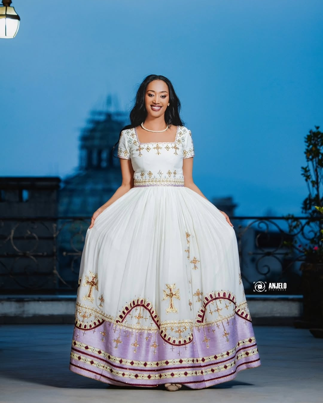 Woman in a white and gold embroidered Eritrean Dress standing on a rooftop at night