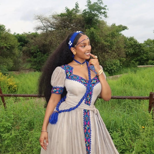 Woman in Habesha Kemis attire with a blue headband and decorative dress standing in a grassy field.