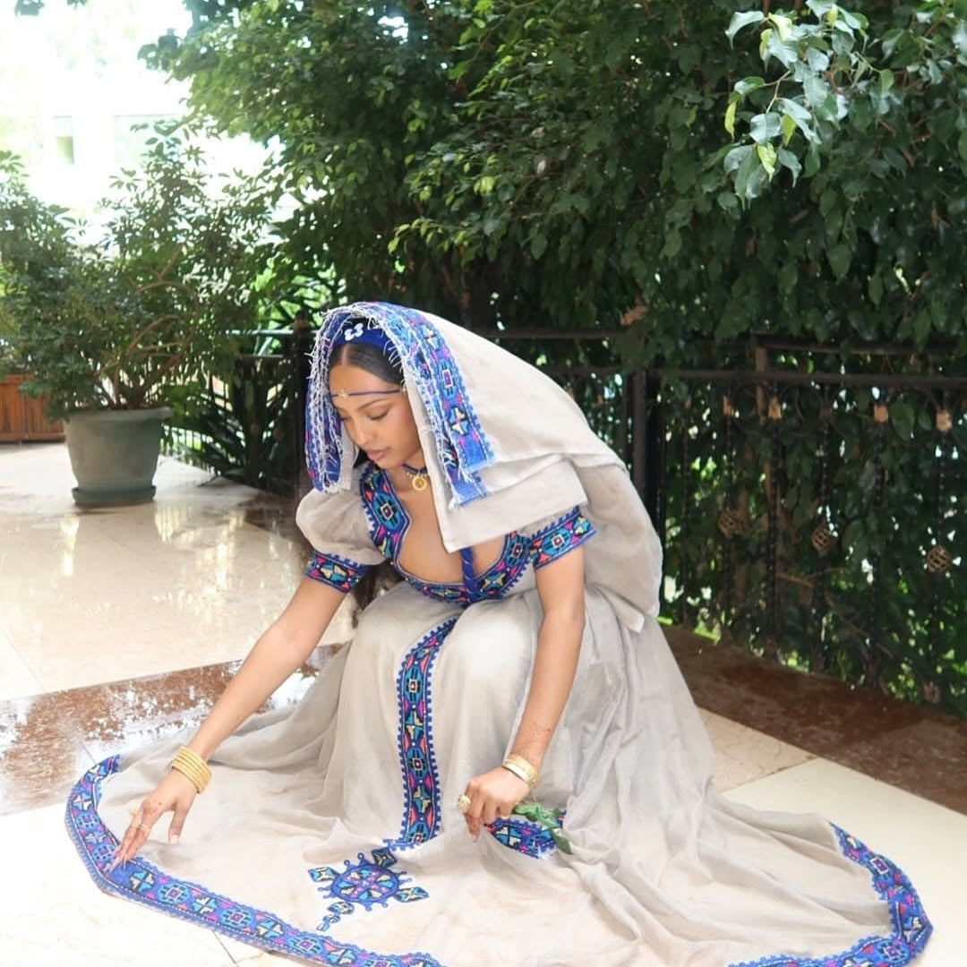 Woman in a Habesha Kemis with blue and white patterns sitting outdoors.