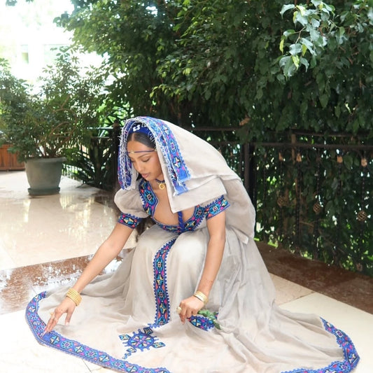Woman in a Habesha Kemis with blue and white patterns sitting outdoors.