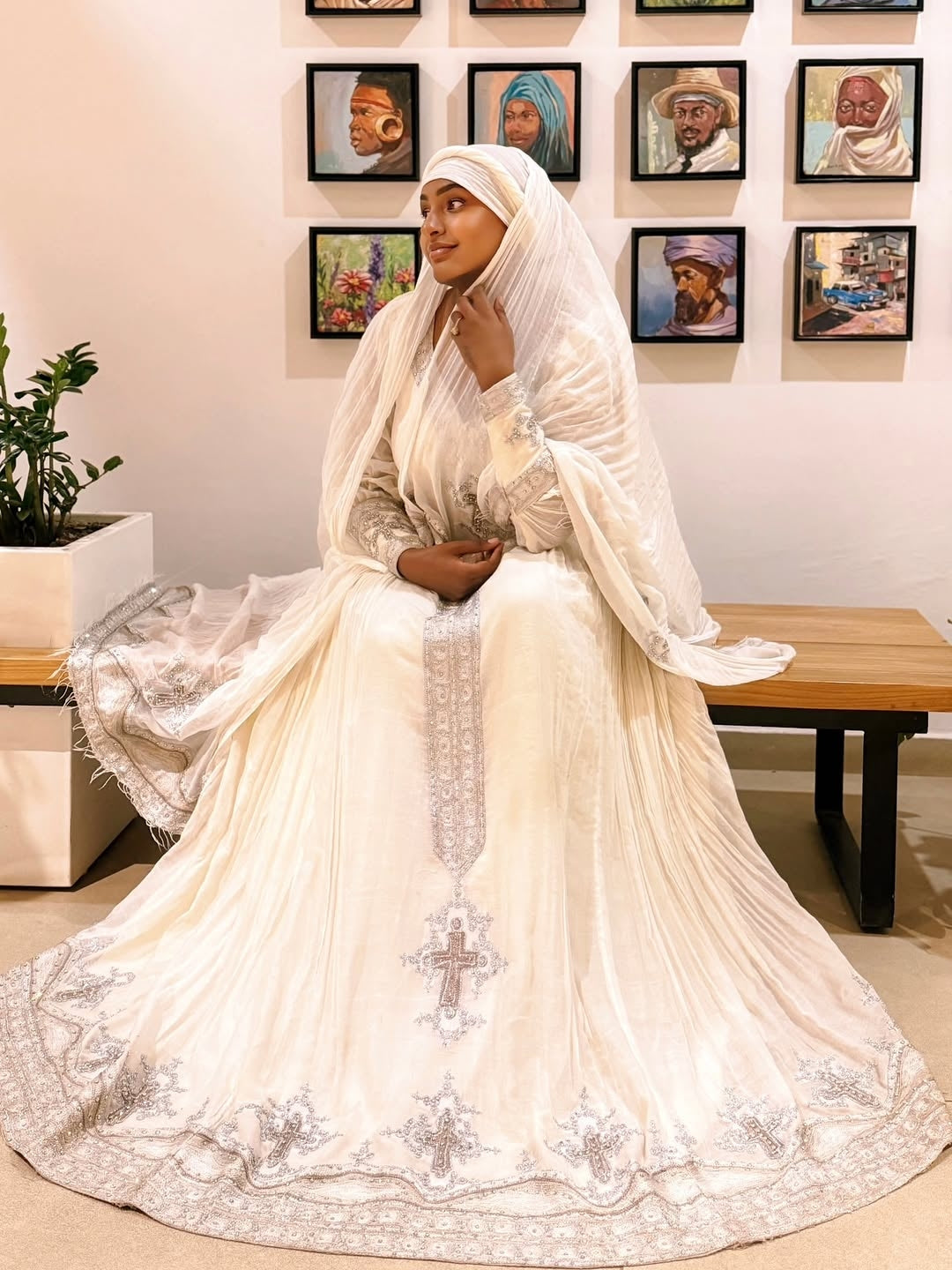 Woman in a white embroidered dress sitting in a room with framed pictures on the wall.