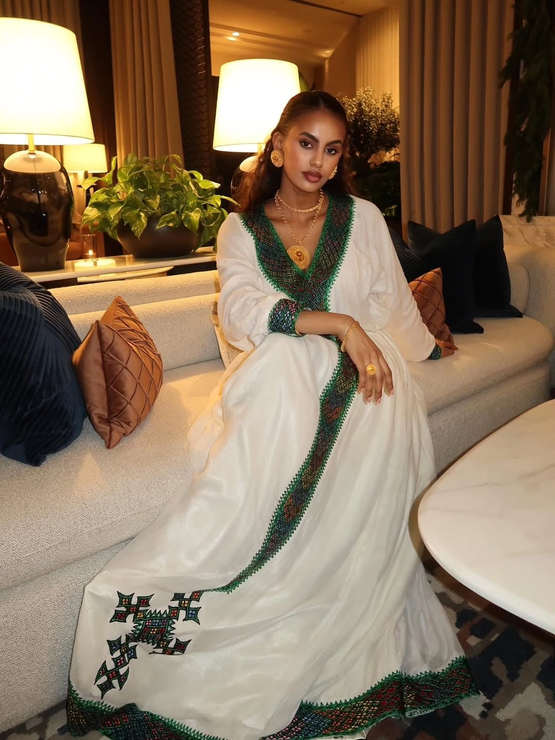 Woman in a white dress with green and red Habesha Libs  sitting on a couch in a decorated room.