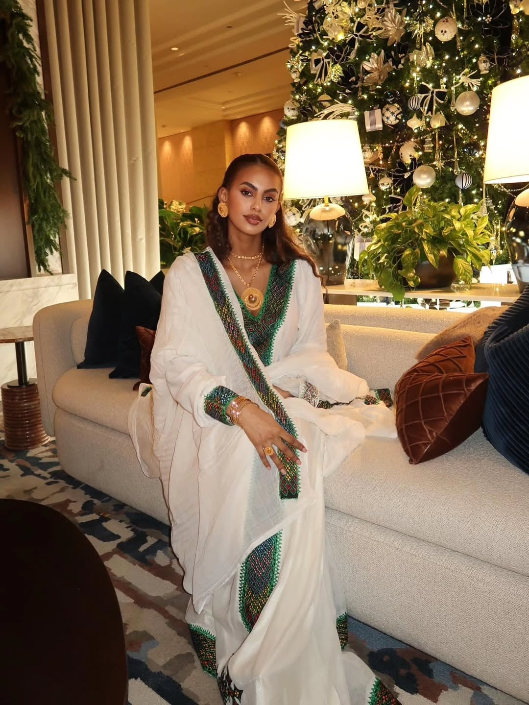 Woman in a white saree with green and gold Habesha Libs  sitting on a couch in a decorated room.