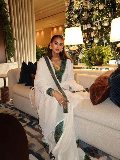 Woman in a white saree with green and gold Habesha Libs  sitting on a couch in a decorated room.
