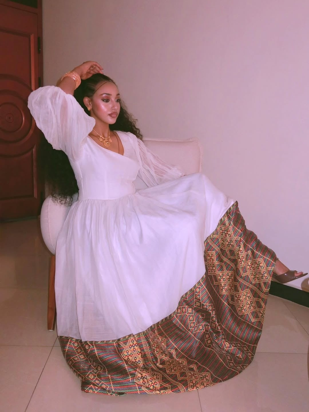 Woman in a Gold Ethiopian Dress dress with a patterned skirt sitting on a chair.