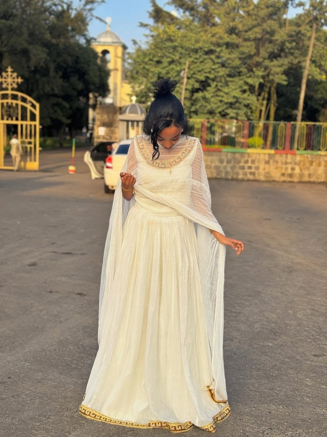 Woman in a white traditional outfit standing on a road with trees and a building in the background