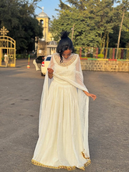 Woman in a white traditional outfit standing on a road with trees and a building in the background